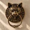 Antique brass cat head towel ring on a sunlit cream wall, its darkened aged finish bringing out the cat's face and Victorian scrollwork. A handmade antique towel ring and gothic bathroom accent, also usable as a cat door knocker, cast in Ubud, Bali.
