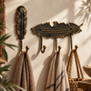 Antique brass finish feather wall hook set of 2: a single feather hook and a 3-hook feather coat rack holding a striped towel, linen towel and woven bag on a sunlit cream wall. Handmade cast brass hooks with a darkened aged patina that highlights the carved feather detail, hand-finished in Ubud, Bali.