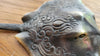 Close-up of an antique brass stingray hook on wood, highlighting the engraved texture and vintage patina from the hook set.