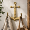 Brass anchor wall hook mounted on a cream wall with a wood frame, holding a striped towel and woven bag, nautical maritime coat hook