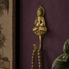 Gold brass sitting buddha wall hook mounted on deep plum textured wall with mala prayer beads hanging from hook and tropical plant beside showing moody spiritual home decor lifestyle styling. Handcrafted sitting buddha coat hook, towel hook and key hook for yoga room decor, meditation space decor, zen home decor and housewarming gift. Made by professional artisans in Bali.