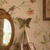 Close-up of single brass butterfly wall hook on vintage floral wallpaper beside ornate gold mirror with soft fabric drape showing intricate wing detail and warm brass finish in whimsical nursery setting. Handcrafted decorative coat hook from set of 4 for nursery room decor, girls room wall decor, baby shower gift and gender reveal gift. Unique new home housewarming gift for butterfly nursery