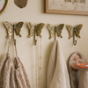 Set of four brass butterfly wall hooks mounted in a row on warm neutral cottagecore wall with linen towels and vintage picture frame showing complete whimsical entryway and nursery styling. Handcrafted decorative coat hook set for nursery room decor, kids room wall decor, baby shower gift and housewarming gift. Unique gender reveal gift and new home gift for whimsical butterfly nursery interiors