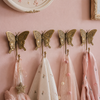 Set of four brass butterfly wall hooks mounted on soft pink nursery wall with delicate pink tulle, spotted fabric and pom pom garland hanging from hooks showing whimsical girls nursery room lifestyle styling. Handcrafted decorative coat hook set for nursery room decor, kids room decor, baby shower gift, gender reveal gift and new home housewarming gift for young families and butterfly nursery lovers