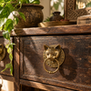 Brass cat head ring pull mounted on the drawer of a dark wooden dresser beside a potted plant and carved decor. A handmade brass cat head drawer pull that also works as a towel ring or door knocker for vintage and dark academia decor, from Bali.