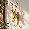 Gold brass deer antler hook on a sunlit cream wall beside a dark wooden frame and green olive branches, casting long antler shadows. A handmade rustic wall hook for hunting cabin decor and woodland decor, hand-finished in Ubud, Bali.