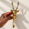 Hand holding the brass deer antler hook against a cream wall, showing its size and the polished gold finish on the stag's head and tall antlers. A handmade rustic wall hook and Christmas gift for deer lovers, hand-cast in Ubud, Bali.
