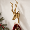 Brass deer Christmas stocking holder in polished gold, mounted on a cream wall beside pine branches and red berries and holding a deep red knit stocking. A handmade stag wall hook with tall antlers, perfect for rustic Christmas and woodland decor, hand-cast in Bali.
