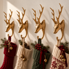 Brass deer Christmas stocking holders set of 4 in polished gold, mounted in a row on a sunlit cream wall and holding red, cream, green and Nordic knit stockings with pine sprigs and bells. Handmade stag wall hooks for a family Christmas mantel display, hand-cast in Bali.
