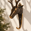 Close-up of the dark antique brass deer head hook beside green plants, showing the darkened stag face, antlers and polished hook tip. A rustic cabin decor stag hook, handmade in Bali.