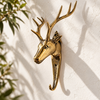 Angled view of the gold brass deer head hook on a cream wall with soft leaf shadows, showing the branching antlers, ears and curved hook. A handmade brass animal hook and antler wall decor piece from Ubud, Bali.