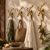 Four brass deer head wall hooks mounted in a row in an entryway, holding a scarf, woven bag, white towel and hanging bells. Shows how the brass deer hook set of 4 creates a rustic cabin decor coat hook wall, handmade in Bali.