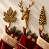 Brass deer, pine tree and maple leaf wall hooks set of 3 in polished gold, mounted on a cream wall and each holding a red and white Christmas stocking with pine sprigs and gift tags. A handmade brass hook set used as Christmas stocking holders for rustic cabin holiday decor, hand-cast in Bali.