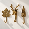 Brass maple leaf hook, deer wall hook with antlers and pine tree hook mounted in a row on a sunlit cream wall with soft shadows. A handmade brass wall hook set of 3 that works as coat hooks or towel hooks all year, cast in Ubud, Bali.