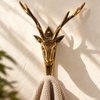 Front view of the gold brass deer head hook holding a beige knit towel, with symmetrical antlers rising above. A handmade towel hook and coat hook for bathrooms, entryways and cabins, cast in Bali.