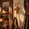Brass deer wall hook in a rustic lodge entryway, mounted beside a wooden post next to a framed stag painting, a lantern and antlers, holding plaid scarves and a leather strap. A handmade rustic cabin decor and lodge decor piece, and a thoughtful hunter gift, cast in Bali.