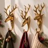 Brass deer wall hooks set of 3 in polished gold, mounted in a row on a cream wall and holding green, red and cream knit Christmas stockings with pine branches, pinecones and bells. Three different stag designs used as Christmas stocking holders for rustic Christmas and woodland decor, handmade in Bali.