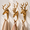 Three brass deer wall hooks in different designs mounted on a sunlit cream wall, holding cream, pink striped and beige towels. A rustic coat hook and towel hook set that works all year after Christmas, a handmade antler hook set for hunting cabin decor, cast in Bali.