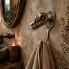 Brass dinosaur skull wall hook mounted in a moody candlelit bathroom with linen towel hanging beside an antique mirror — gothic dark academia towel hook handmade in Ubud Bali
