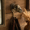 Solid brass dinosaur skull wall hook mounted on an ornate rustic wood wall with dark coat and dried pampas grass hanging — gothic dark academia wall hook handmade in Ubud Bali