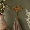 Gold brass fan leaf wall hook mounted on warm olive green boho wall with large tropical plant and soft linen towel hanging showing nature-inspired lifestyle styling. Handcrafted leaf shaped plant hanger hook, towel hook and coat hook for boho home decor, housewarming gift and botanical nature-inspired entryway and bathroom wall decor. Available in gold and dark antique finish