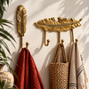 Set of 2 gold brass feather wall hooks: one single feather hook and one 3-hook feather coat rack, mounted on a warm cream wall holding a rust towel, striped towel and woven straw bag. Handmade cast brass boho entryway and bathroom hooks, hand-finished by artisans in Ubud, Bali, shown in soft natural light with green plants.