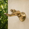 Angled view of a solid brass fingering hand wall hook mounted near greenery, showing the sculptural hand design.