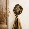 Antique brass Ganesha wall hook mounted on warm white textured wall with decorative tassel, hanging keys and dried botanical grass arrangement beside rustic wooden frame showing boho entryway lifestyle styling. Handcrafted Buddhist elephant wall hook for boho home decor, yoga room decor, meditation space and housewarming gift. Created with reverence by professional Hindu artisans in Bali