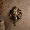 Antique brass Ganesha wall hook mounted on dark warm brown textured wall showing rich patina finish, intricate ornate crown, detailed elephant face and trunk hook. Handcrafted Buddhist elephant wall hook for boho home decor, yoga room decor, meditation space and unique housewarming gift. Created respectfully by professional Hindu artisans in Bali