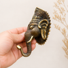 Hand holding antique brass Ganesha wall hook beside dried pampas grass showing rich dark patina finish, intricate Ganesha crown and detailed elephant trunk hook. Handcrafted Buddhist elephant wall hook for boho home decor, yoga room decor, meditation decor and unique housewarming gift. Created with reverence by professional Hindu artisans in Bali