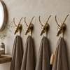 Four solid brass impala skull wall hooks mounted in a row on a neutral wall with grey towels hanging showing complete skull coat hanger set — gothic dark academia decor handmade in Ubud Bali