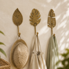 Set of three brass leaf wall hooks — Monstera, Fern, and Spade Leaf — holding a woven hat, tote bag, and scarf on a boho wall