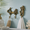 Two brass mermaid wall hooks mounted on soft teal coastal bathroom wall with white fluffy towel and coral reef decoration showing complete beach house bathroom styling. Handcrafted gold brass mermaid double hook and single hook for coastal nautical home decor, beach house bathroom and housewarming gift for ocean lovers