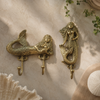 Two brass mermaid wall hooks styled on warm neutral surface beside large seashell and macrame tassel showing coastal nautical lifestyle detail. Handcrafted gold brass mermaid double hook and single hook set for beach house decor, coastal bathroom styling and unique housewarming gift and new home gift for ocean lovers