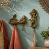 Brass mermaid wall hook set mounted on teal coastal wall with coral pink towel and macrame tassel hanging showing beach house bathroom lifestyle styling. Handcrafted gold brass mermaid double hook and single hook for coastal nautical home decor, beach house bathroom decor and housewarming gift for ocean lovers