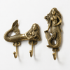 Brass mermaid wall hook set on white background showing double hook mermaid and single hook mermaid in warm gold finish with full sculptural mermaid detail, flowing hair and scaled tail. Handcrafted coastal nautical home decor, decorative coat hook and towel hook set. Unique beach house housewarming gift for mermaid decor lovers