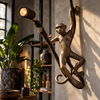 Antique brass monkey lamp mounted on a stone wall in a tropical living room beside leafy plants, shelves and dark pottery, its spotlight casting a warm glow. A plug-in hanging monkey wall sconce that adds jungle decor and safari style to an eclectic home, handmade in Bali.