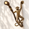 Angled view of the antique brass monkey sconce on a cream wall with soft leaf shadows, showing the depth of the hanging monkey, the round wall plate and the glowing spotlight. A handmade plug-in monkey wall lamp with 6 ft cord and US plug, cast in Bali.