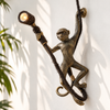Brass monkey wall lamp in antique brass on a sunlit white wall with palm leaf shadows, the hanging monkey gripping a woven vine and holding a glowing spotlight. A handmade plug-in hanging monkey sconce with US plug and bulb included (vine shown for styling, not included), cast in Bali.