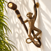 Brass monkey wall lamp switched on, its spotlight glowing as the hanging monkey grips a styling vine on a sunlit wall with tropical leaf shadows. A handmade plug-in monkey sconce in antique brass, ready to use with the included bulb and US plug.
