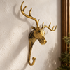 Brass moose wall hook on a cream wall beside a dark wooden frame and pine branches, casting soft antler shadows. A handmade rustic cabin decor piece and whimsical Christmas stocking holder in polished gold, hand-cast in Ubud, Bali.