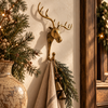 Brass moose hook holding a linen towel and a brass bell beside pine branches, a vase of greenery and a wooden mirror frame. Whimsical Christmas decor that works as a towel hanger or stocking holder, a handmade rustic cabin decor piece from Bali.