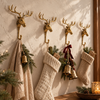 Four brass moose hooks mounted in a row above a wooden mantel, holding cream knit Christmas stockings, a towel and brass bells with pine sprigs. A whimsical Christmas stocking holder display and festive mantel decor, a perfect moose lover gift, handmade in Bali.