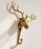 Brass moose wall hook in shiny polished gold on a cream wall, showing the moose head in profile with tall branching antlers and a curved hook below. A handmade brass animal hook and whimsical Christmas stocking holder, hand-cast in Ubud, Bali.