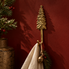 Brass pine tree wall hook in polished gold, mounted on a deep red wall beside Christmas garland and holding a cream stocking with a jingle bell. A handmade Christmas tree hook and stocking holder that adds rustic cabin charm to holiday wall decor, hand-cast in Bali.