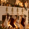 Four brass pine tree hooks mounted along a white fireplace mantel, each holding a Christmas stocking beneath garland, candles and a crackling fire. Shows how the brass Christmas tree hooks work as stocking holders for the whole family, handmade in Bali.