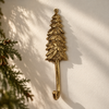 Gold brass pine tree wall hook on a sunlit cream wall with soft evergreen branch shadows, showing the textured tree branches and curved hook. A handmade evergreen coat hook and tree wall hook for cabins, entryways and woodland-style homes, cast in Bali.