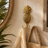 Brass pineapple double wall hook polished gold mounted on warm textured wall beside rattan mirror holding woven bags and linen towel with tropical plants – pineapple wall hook coat hook towel hook – tropical boho beach house coastal home decor – housewarming gift – handcrafted solid brass made in Bali by UbudGlassandBrass

