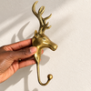 Brass reindeer wall hook held in a hand against a cream wall, showing the stag's head in profile, the branching antlers and the long curved hook with ball tip. A handmade brass deer hook and coat hook from Ubud, Bali.