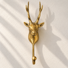 Straight-on front view of the brass reindeer wall hook on a cream wall with soft leaf shadows, showing the symmetrical antlers, the stag's face and the hook with ball tip. A handmade Christmas stocking holder and towel hook, cast in Bali.